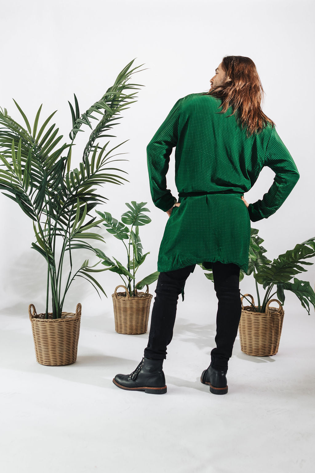 a man in a green robe and black pants standing next to two potted plants